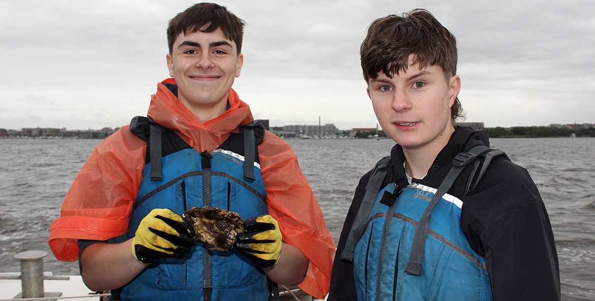 Two boys wearing blue life vests stand with the water behind them, one holding a fresh oyster.